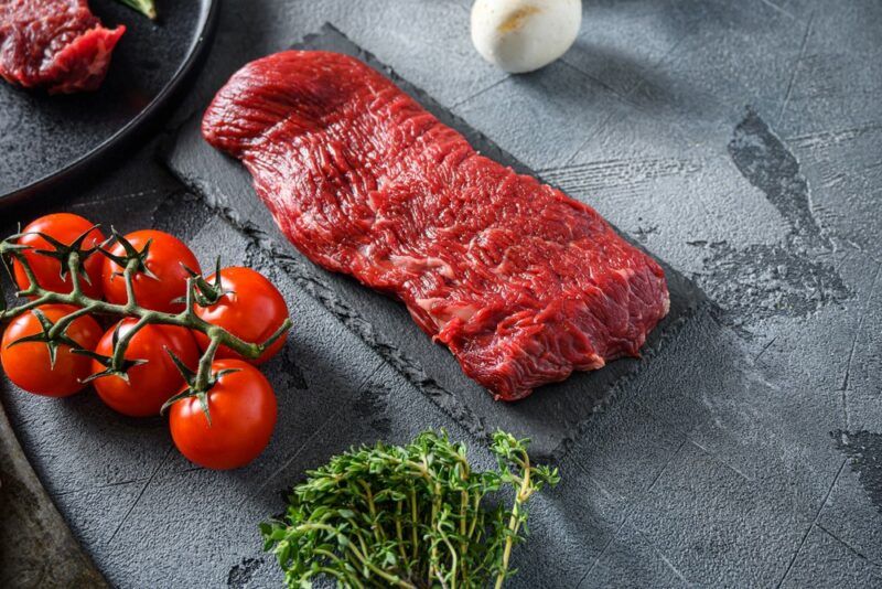 A piece of raw sirloin tip side steak on a table next to cherry tomatoes and fresh herbs