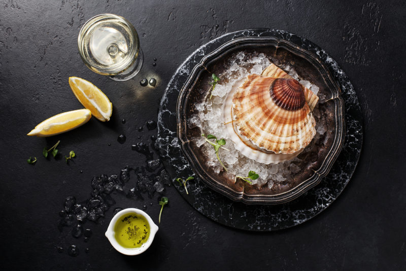 A single scallop in its shell in a black dish with ice, next to a glass of wine with lemon wedges