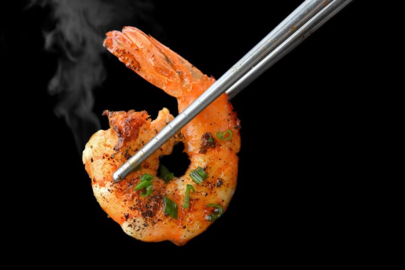 A pair of chopsticks holding up a single cooked and steaming shrimp