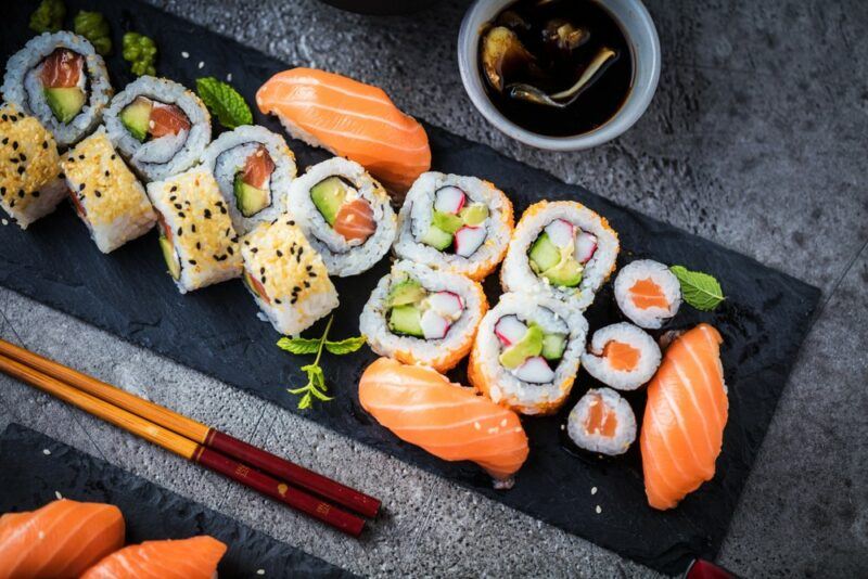 A black slate with many pieces of fresh sushi, some chopsticks, and soy sauce