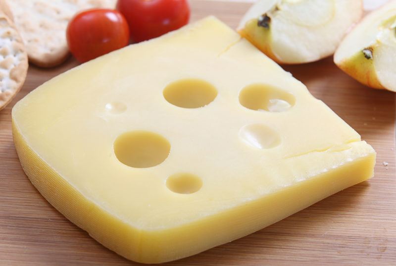 A piece of jarlsberg cheese with tomatoes and crackers in the background