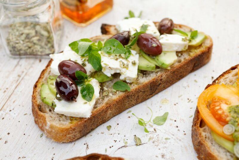 A slice of sourdough that has been topped with avocado, cheese, and olives