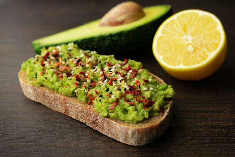 A slice of sourdough with mashed avocado, chili pepper, and lemon juice, with an avocado and half a lemon in the background