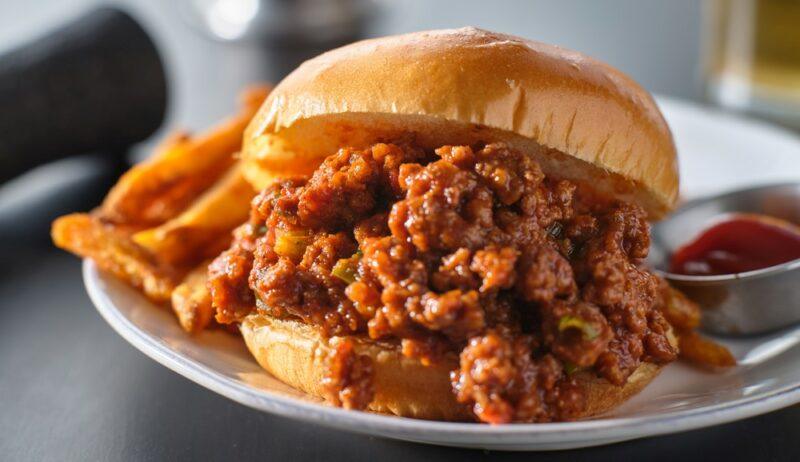 A white plate containing a large sloppy joe with fries