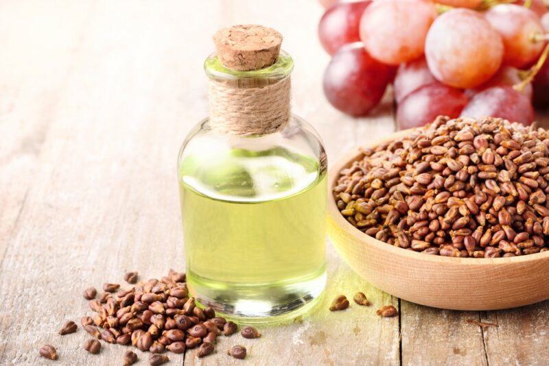 A wooden table with a bottle of grapeseed oil, next to some grape seeds, a bowl of more grape seeds, and some fresh grapes