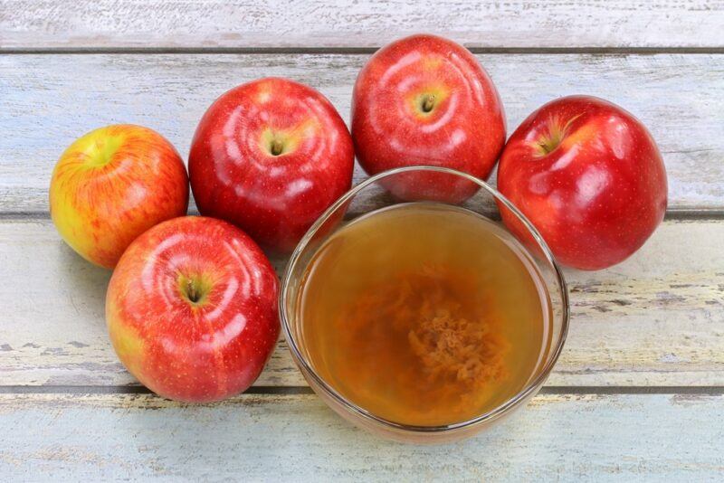 A small bowl of apple cider vinegar with the mother, surrounded by red apples on a white wooden table