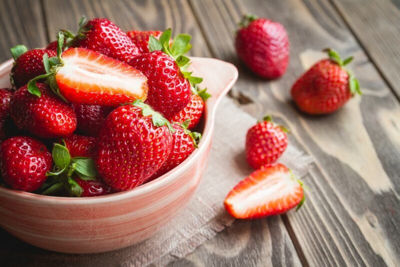 A dish filled with fresh strawberries, one that's been cut in half, with a few more strawberries scattered on a table