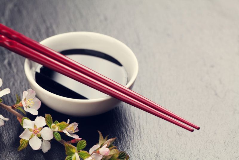 A small white bowl that contains soy sauce, with two chopsticks next to white flowers