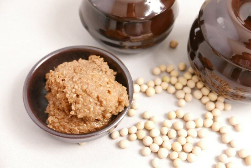 A white table top with a few brown containers, a small brown dish of soybean paste, and many soybeans scattered on the table itself.