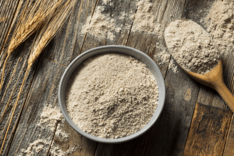 A small metal bowl of whole wheat flour, next to a wooden spoon of the flour and more flour scattered on the table. There are also wheat stalks on the table.