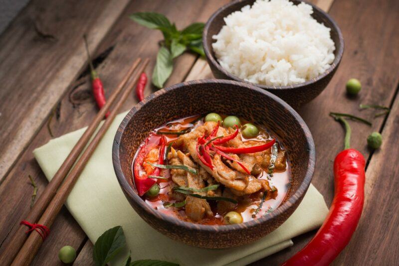 Two small brown bowls, one containing a spicy meal and the other containing freshly cooked rice. There is a pair of chopsticks, a fresh pepper, and some other ingredients on the table.