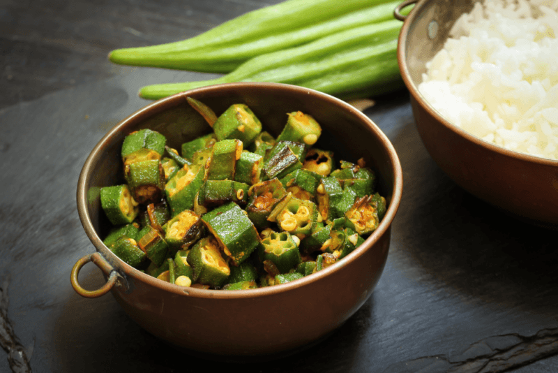 A dark brown bowl with fried okra, next to some fresh okra and a bowl of rice