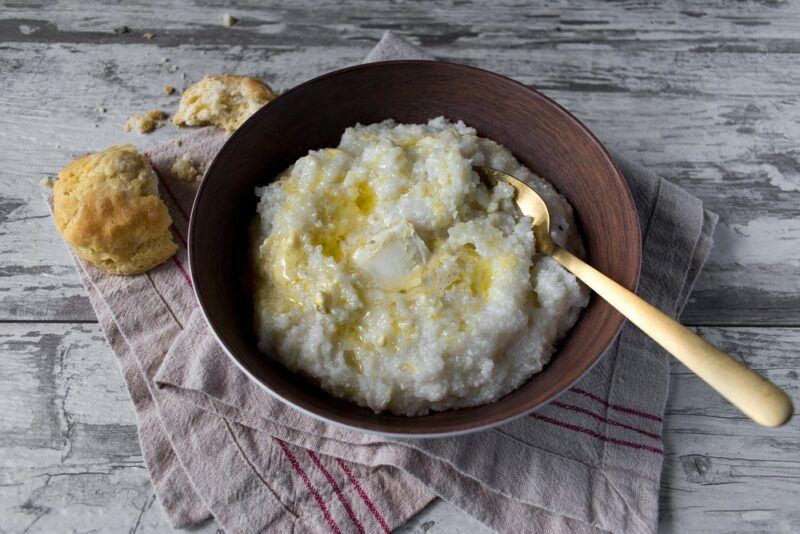 A brown bowl with buttery homemade grits and a spoon, on top of a cloth