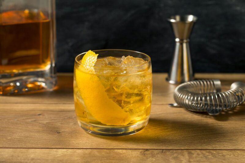 A small glass containing a rusty nail cocktail with plenty of ice and a lemon twist, next to a bottle of Scotch whiskey, a jigger, and a strainer