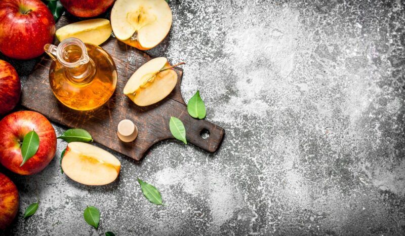 A small jar of apple cider vinegar on a wooden tray next to apples