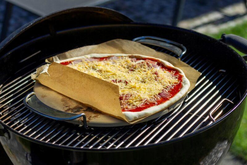 A grill with a pizza stone with handles, plus a pizza with red sauce and cheese