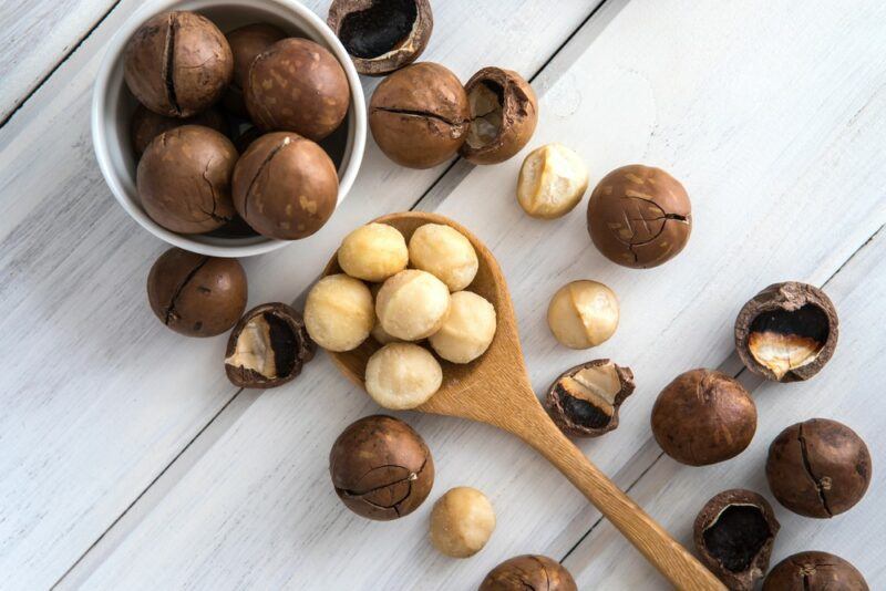 A white wooden table with a spoonful of shelled macadamia nuts, a bowl of more macadamia nuts, and some macadamia nuts scattered across the table