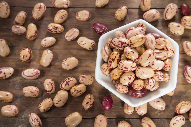 Many pinto beans scattered on a wooden table, surrounding a white dish of pinto beans