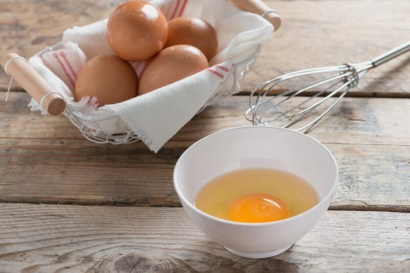 A small white bowl containing a raw egg, next to a basket of eggs