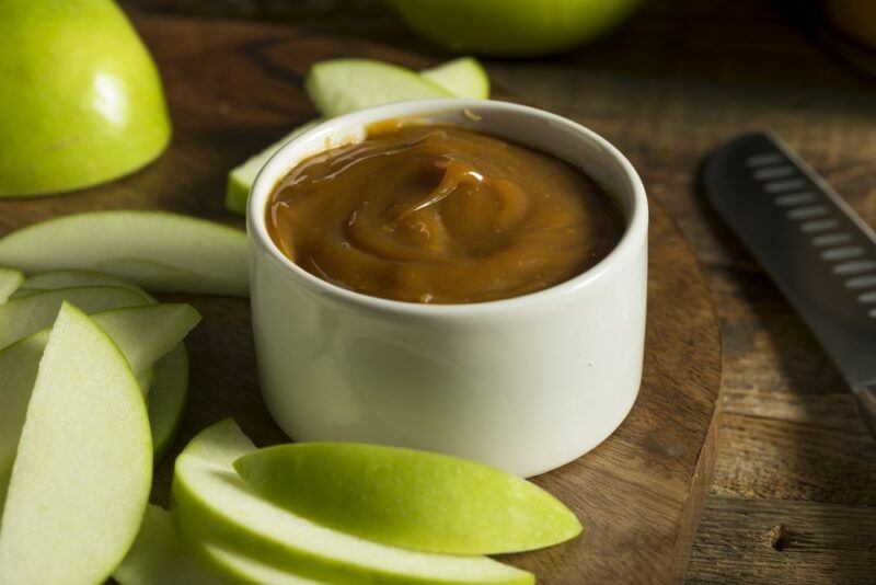 A small white dish of caramel sauce on a brown table, next to bright green apple slices