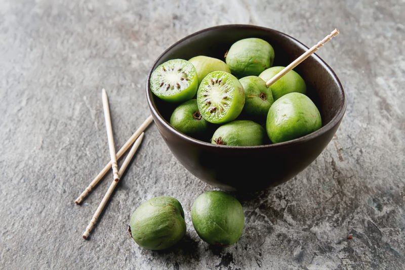 A small black bowl on a table with kiwiberries. Some are whole, others have been sliced. One has been skewered with a toothpick. A few more berries and toothpicks are on the table