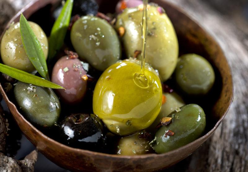 A small brown bowl that contains fresh olives with olive oil being drizzled over them