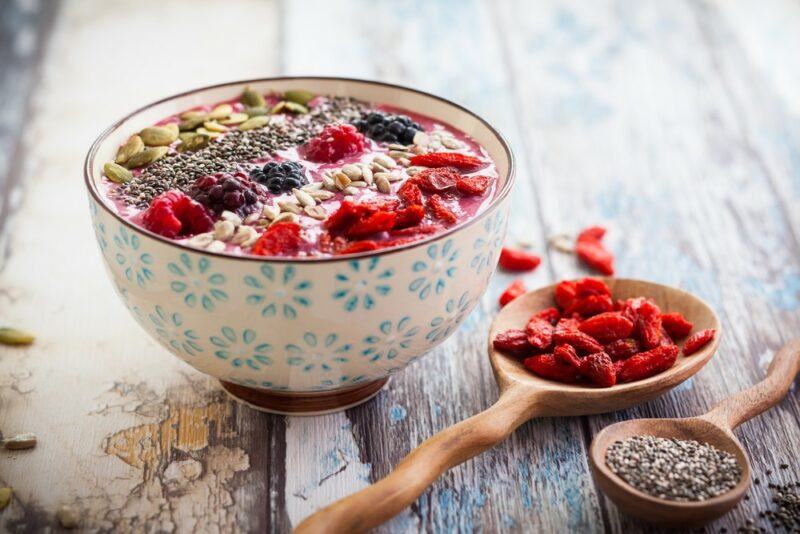 A smoothie bowl topped with various add-ons, including seeds, goji berries, and berries, next to a spoon filled with goji berries