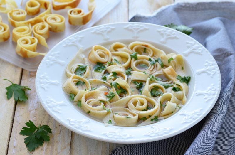 A white bowl with a soup that uses thin slices of pancake. There are more rolled up pancake strips behind the bowl