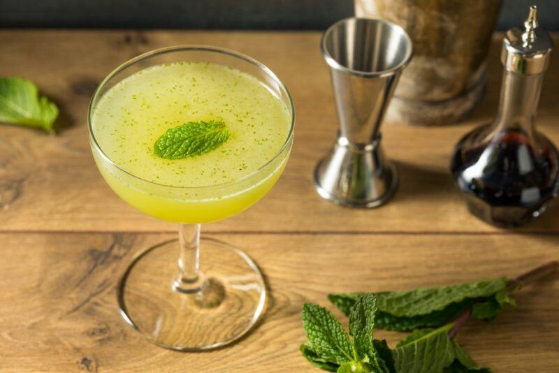 A wooden board or table with a green Southside cocktail, next to bar tools and some leaves