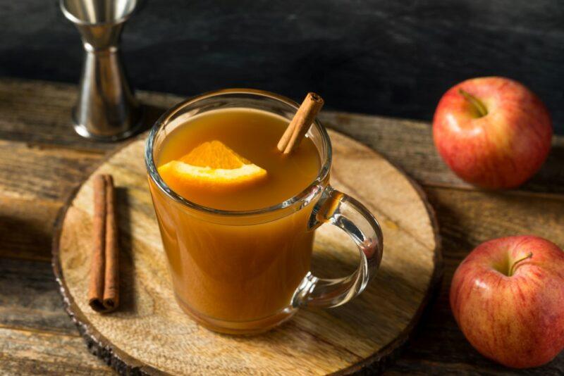 A wooden board with a spiced winter blizzard cocktail next to a cinnamon stick and two apples