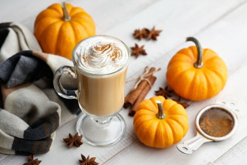 A glass mug containing a spiced pumpkin latte that has been spiced with rum, with pumpkins, spices, and other decorations on the table