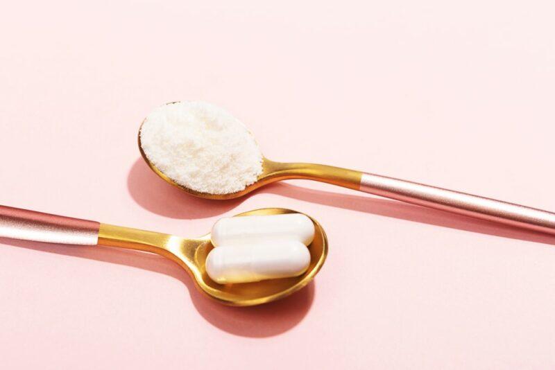 A light pink background with two metallic spoons. One is filled with collagen powder, while the other has collagen tablets instead