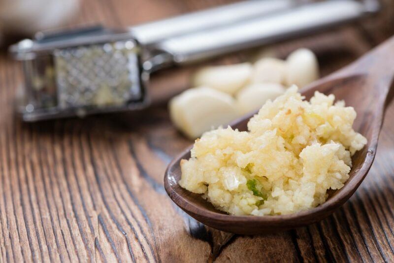 A brown bowl of crushed garlic, in front of a garlic press on a wooden table