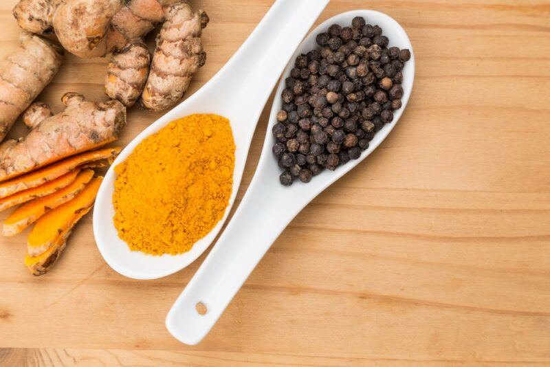 A wooden table with a white spoon full of peppercorns and one of turmeric powder, next to turmeric roots