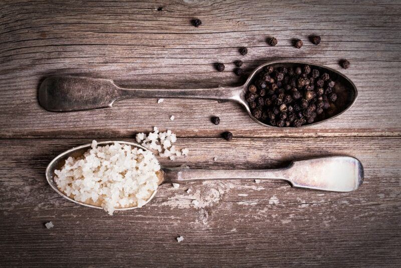 A wooden table with stainless steel spoons. One contains black peppercorns and the other has rock salt