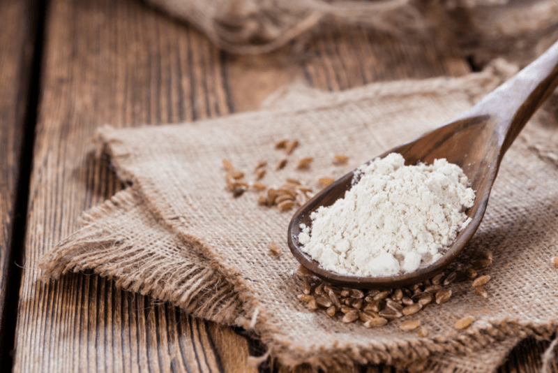 A wooden table with burlap sacks, spelt grains, and a wooden spoon of spelt flour.