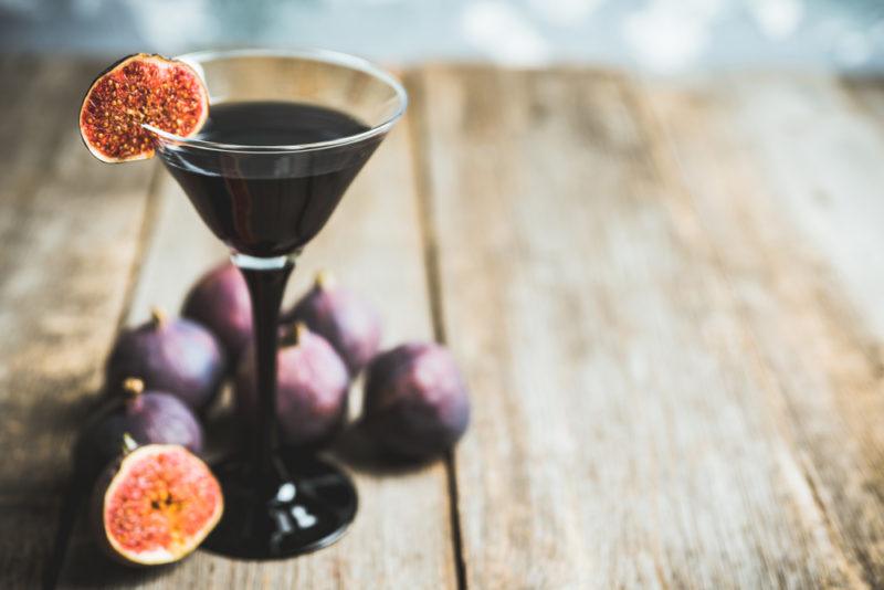 A black cocktail made with squid ink and garnished with a fig on a table with many other figs