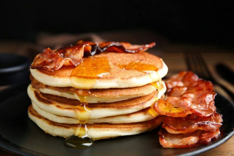 A stack of pancakes on a black dish, with plenty of bacon and maple syrup