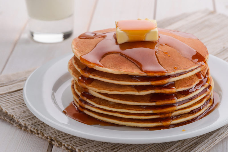 A stack of pancakes on a white plate with a knob of butter on top and maple syrup running down the sides