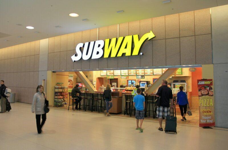 A Subway store in an airport with travelers mingling in front