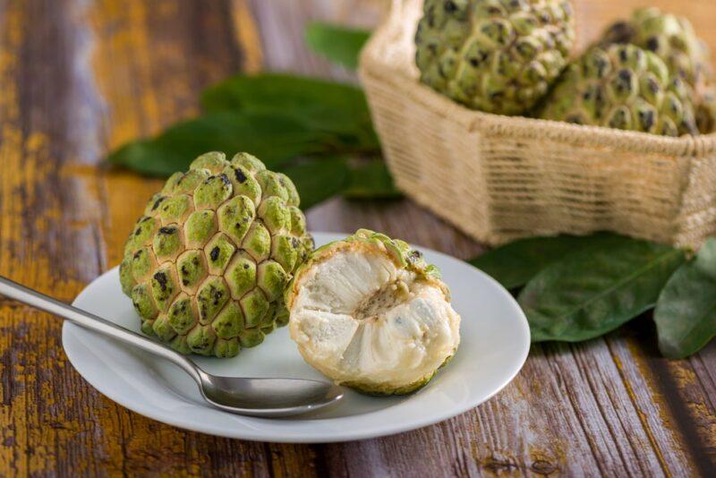 A white plate with a whole and part of a sugar apple and a spoon, with a basket of sugar apples in the background