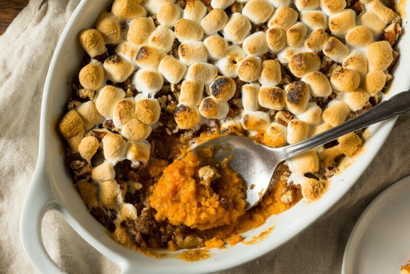 A white dish containing a sweet potato casserole that has been topped with marshmallows and is being delved into with a spoon