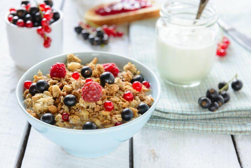 A wooden table with a bowl of cereal and fruit, plus container of berries and milk in the background