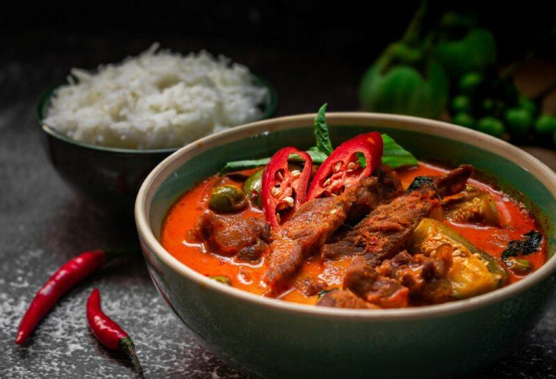 A large bowl containing a spicy curry, with a black bowl of rice and some chili peppers in the background.