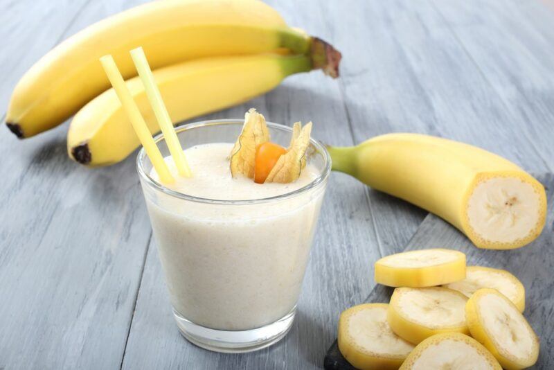 Fresh bananas behind a glass containing a banana smoothie and some banana slices.