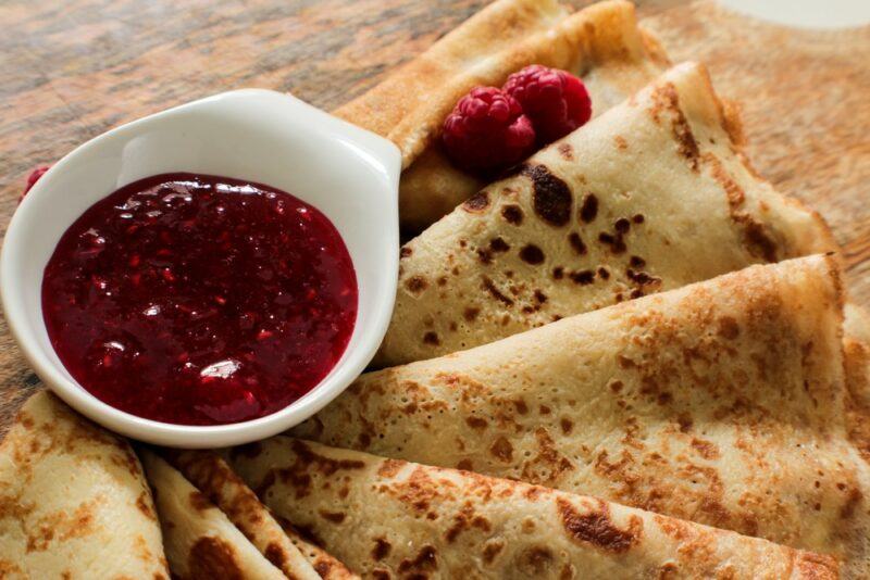 A wooden table with thinly sliced blini pancakes, next to a container of jam