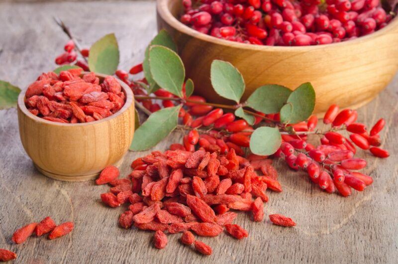 A small bowl filled between goji berries, next to a larger bowl of the berries and more berries on the table