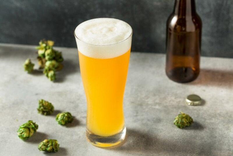 A gray table with a large pint glass of IPA, surrounded by hops, with a single empty bottle in the background