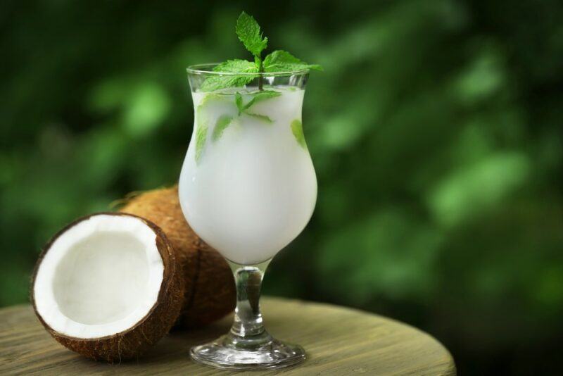 A hurricane glass that contains a White Claw pina colada next to two coconut halves, with out of focus leaves in the background