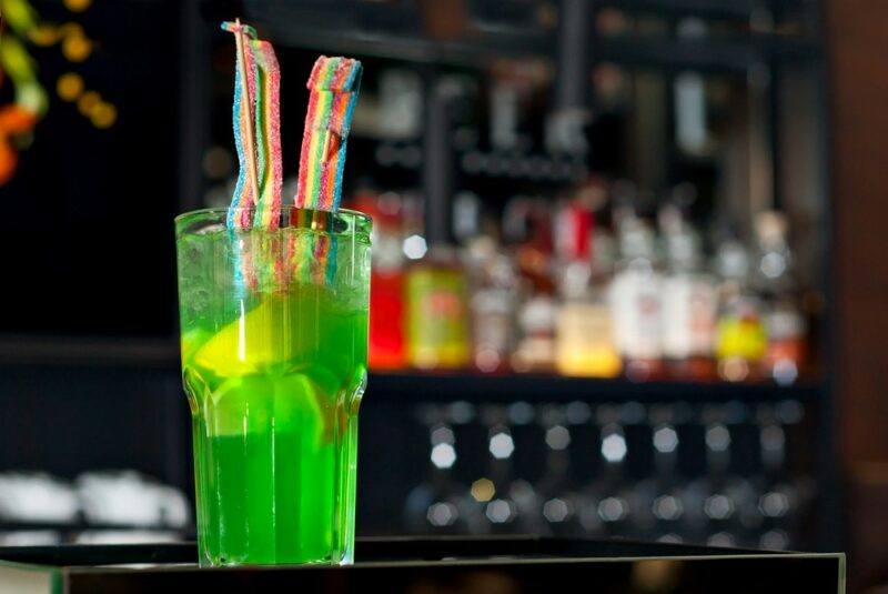 A tall glass containing a bright green nuclear hulk cocktail with lemon and two candy strips as a garnish, in front of an out of focus bar with many bottles